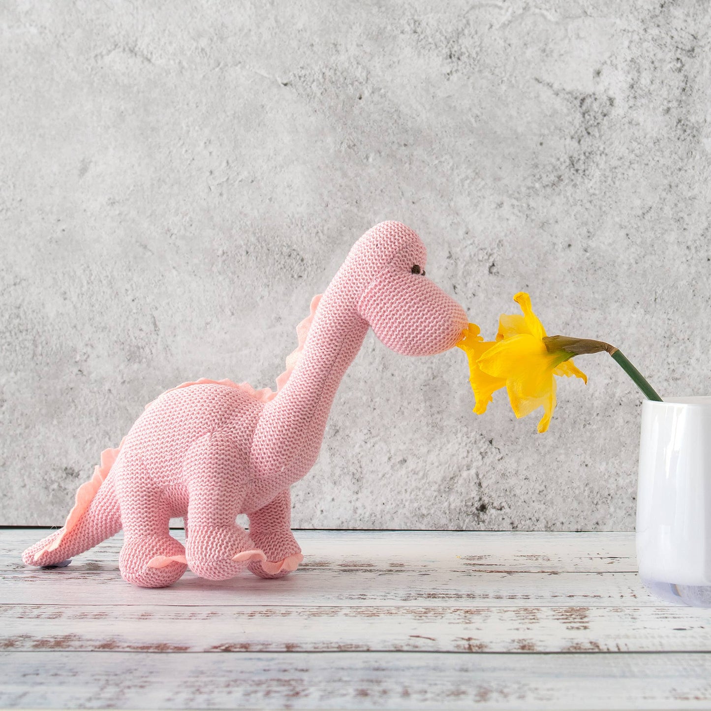 Knitted pink dinosaur toy interacting with a yellow flower on a wooden surface with a concrete wall background.