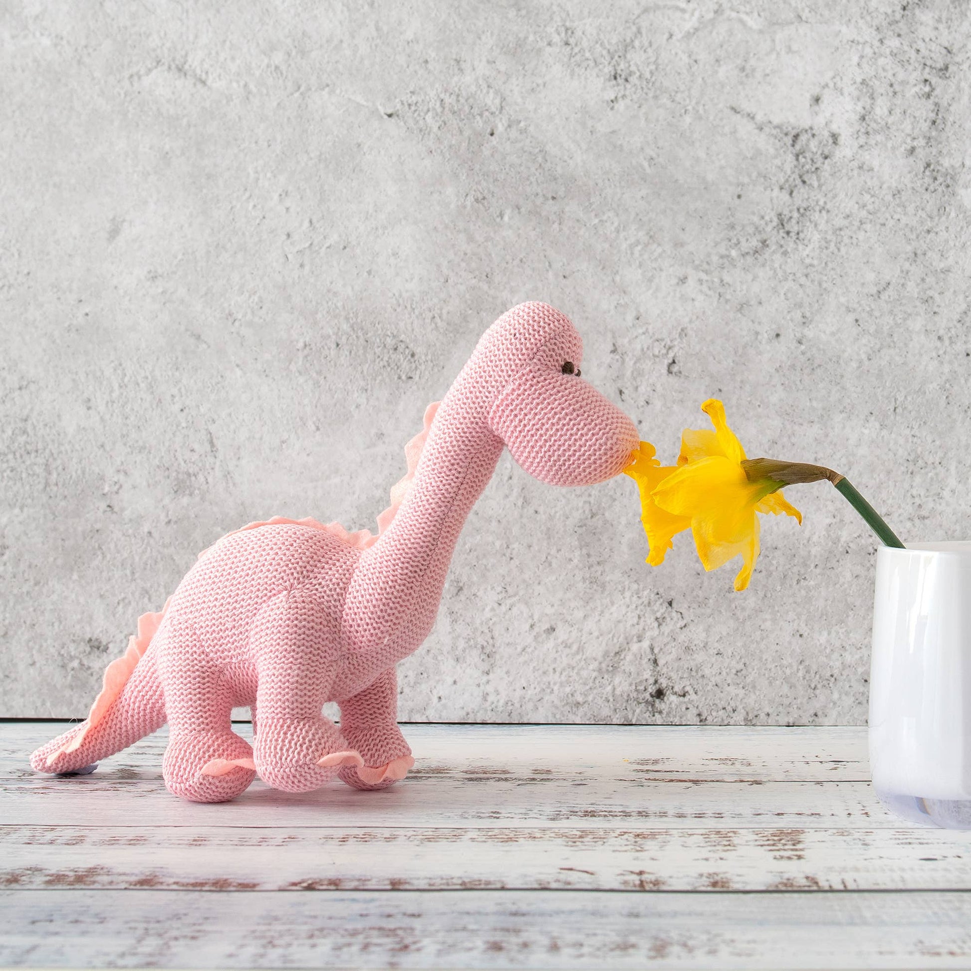 Knitted pink dinosaur toy interacting with a yellow flower on a wooden surface with a concrete wall background.