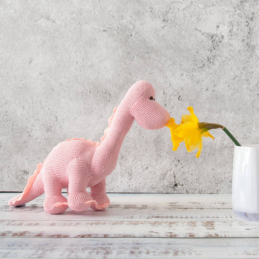 Knitted pink dinosaur toy interacting with a yellow flower on a wooden surface with a concrete wall background.