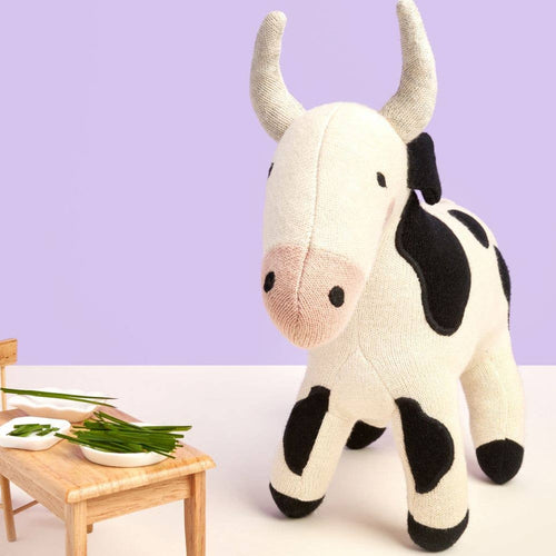 Stuffed cow toy standing next to a small wooden table with a chair and plates on a light purple background
