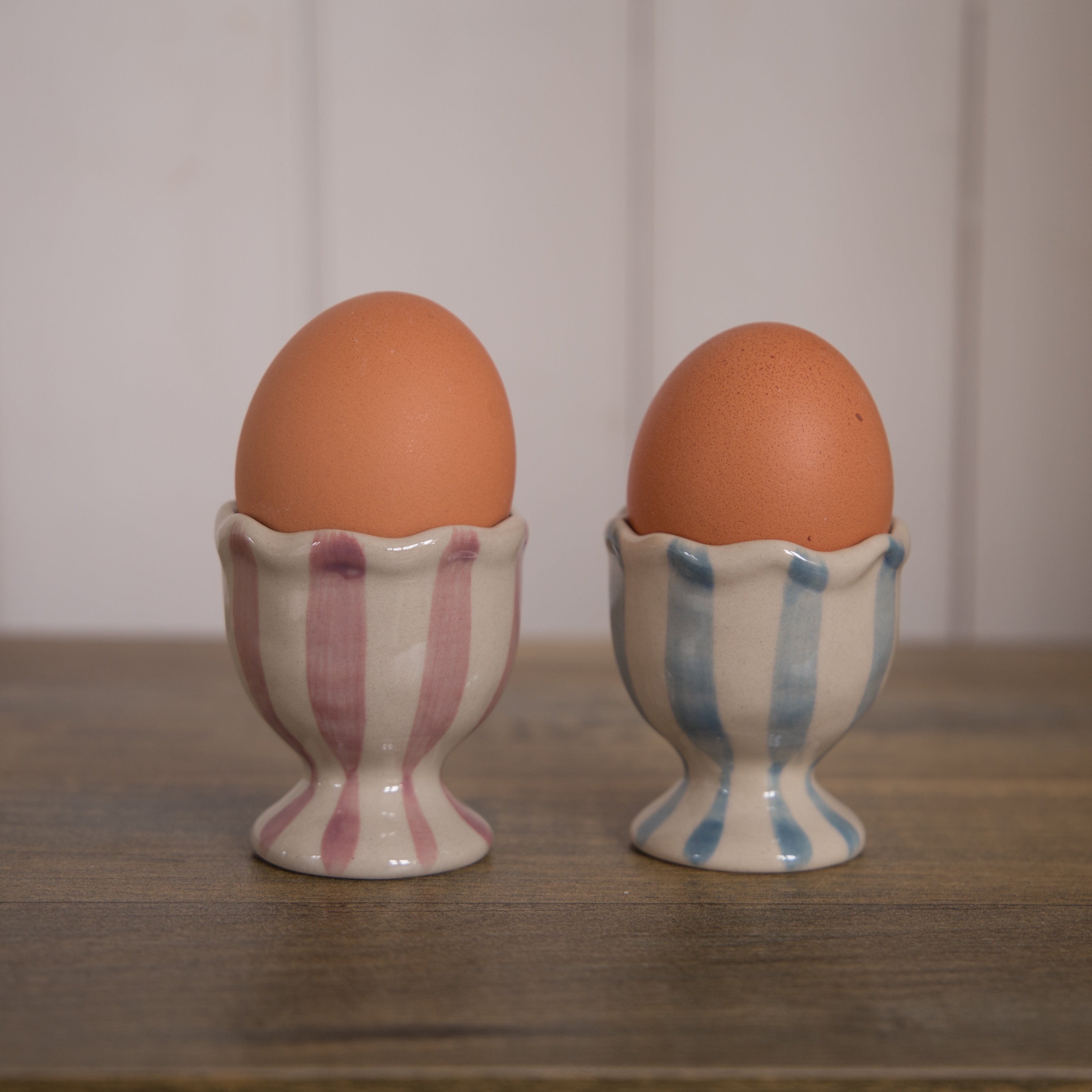 Bloomingville Willa Striped Egg Cups, one in pink and white stripe and one in blue and white stripe.  Both have a ruffle edge and are displayed on a table with a white wall background.