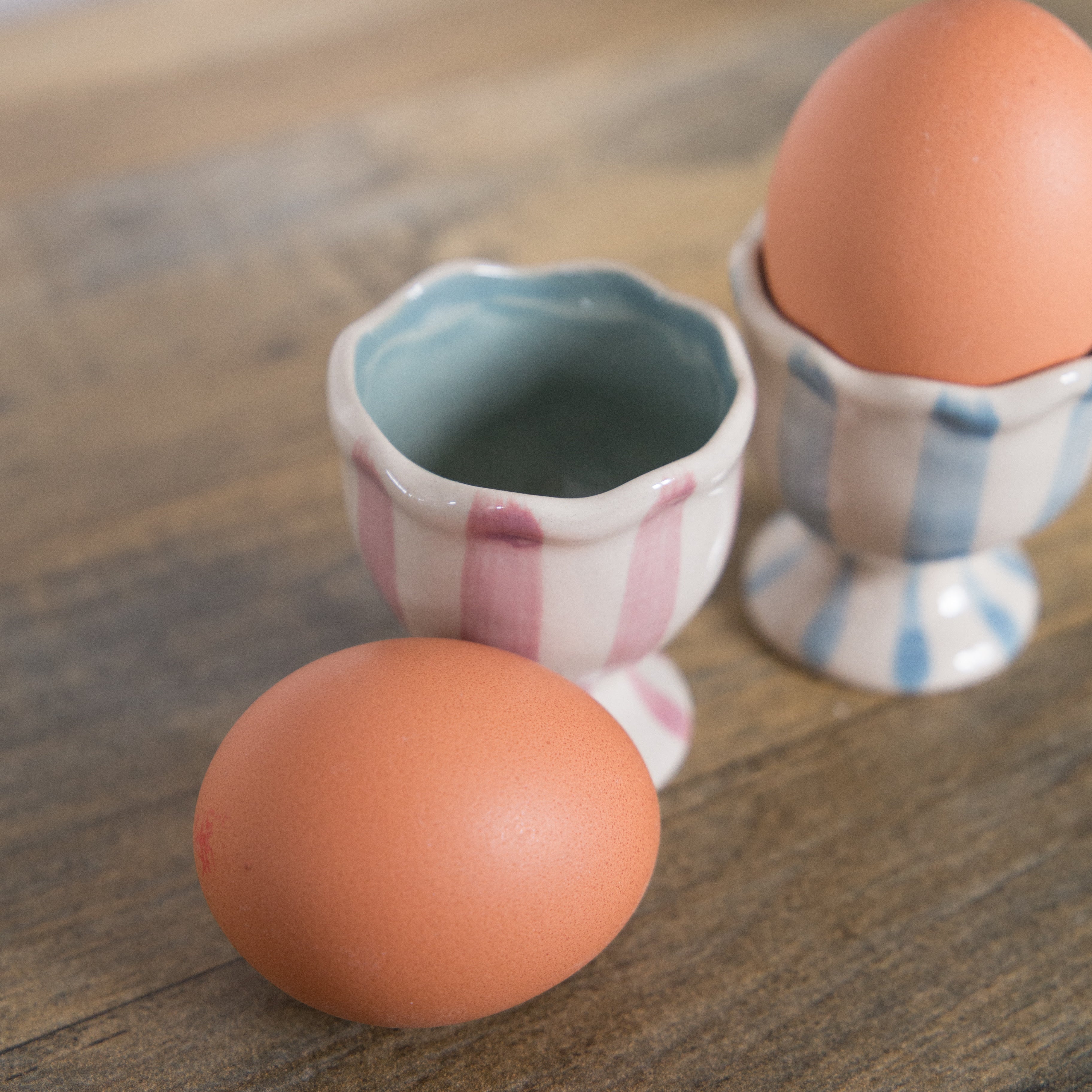 Bloomingville Willa Striped Egg Cups, one in pink and white stripe and one in blue and white stripe.  Both have a ruffle edge and are displayed on a table with a white wall background. The blue and white striped egg cup has an egg placed in it and the pink and white striped one has an egg placed beside it on the table.
