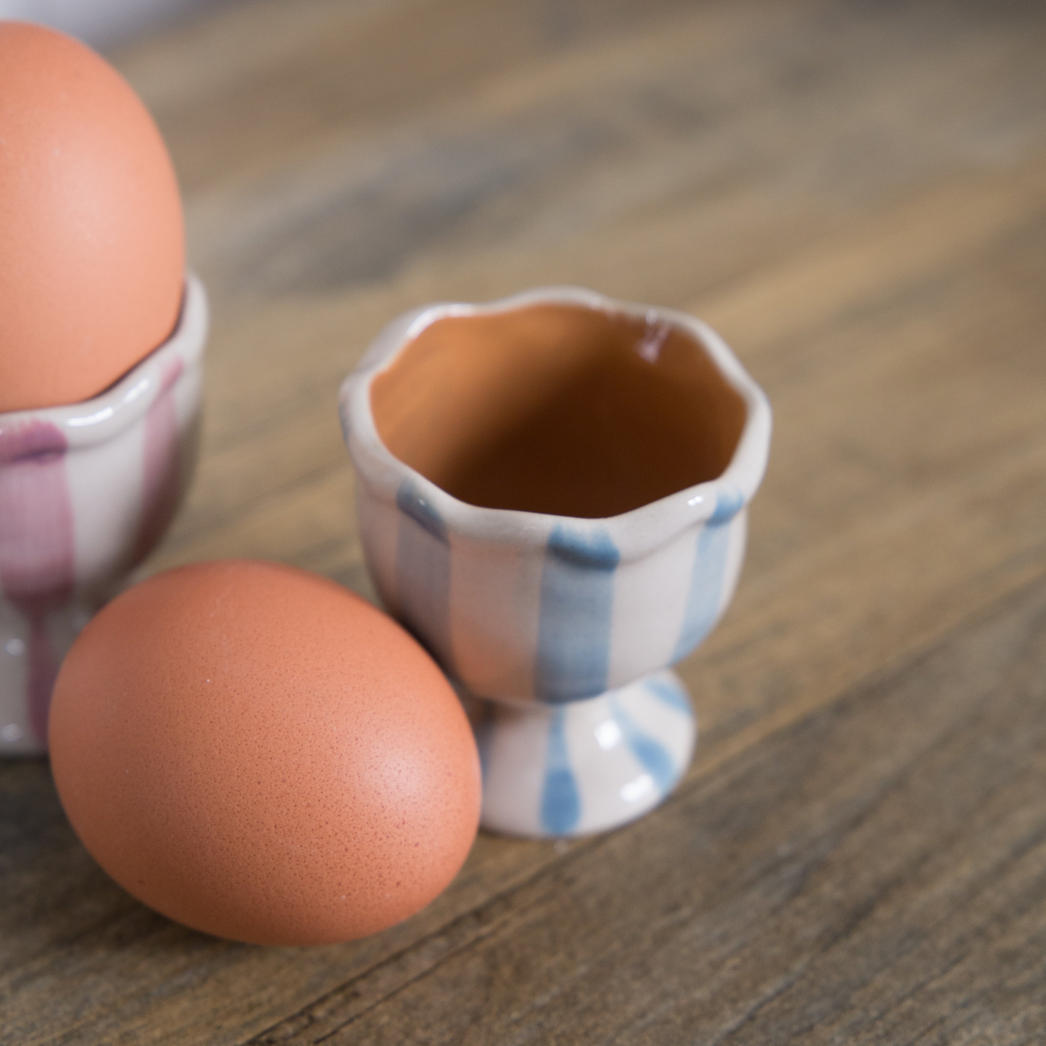 Bloomingville Willa Striped Egg Cups, one in pink and white stripe and one in blue and white stripe.  Both have a ruffle edge and are displayed on a table with a white wall background.  