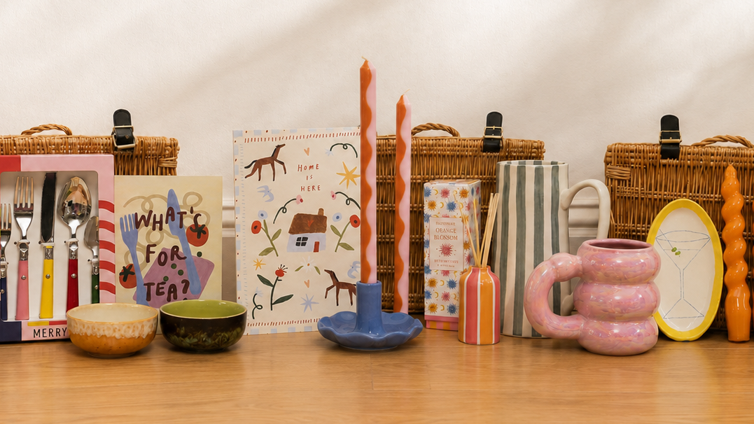 A curated collection of colorful home and dining accessories arranged on a light wood floor in front of a cream wall with soft diagonal shadows. The set includes wicker picnic baskets, a yellow tiered candle holder, patterned cutlery, illustrated cards, tall orange taper candles, a blue candlestick holder, a pink wavy mug, orange spiral coasters, striped and patterned boxes, and a yellow-rimmed trinket dish.