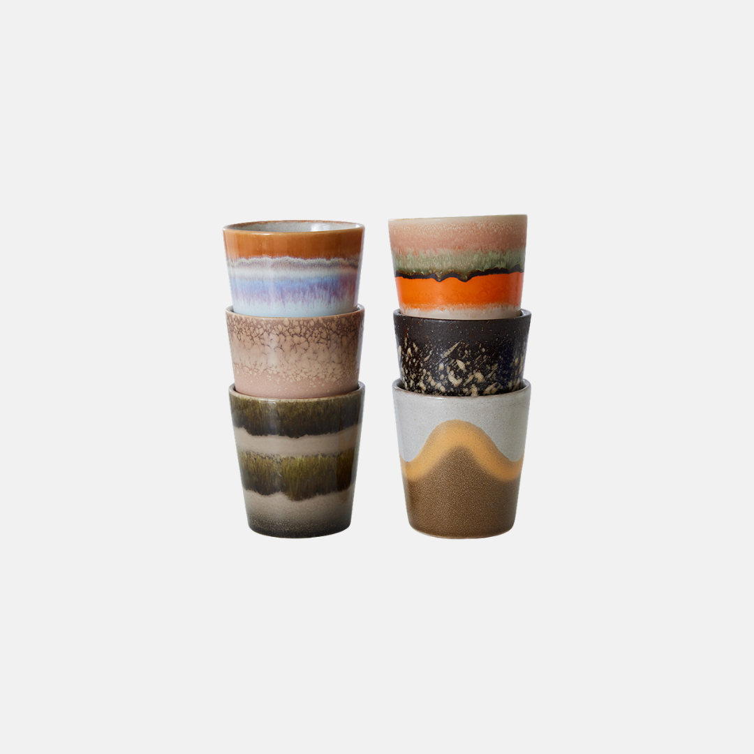 Two stacks of small ceramic cups with abstract earthy patterns in beige, brown, orange, black, and muted green, set against a plain light gray background.