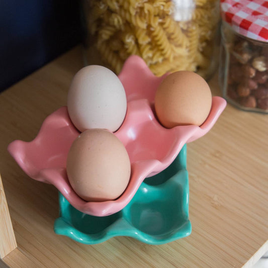 Three eggs in a pink and green egg holder on a wooden surface with jars of pasta and nuts in the background.
