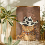 Framed artwork of a leopard wearing a cowboy hat with star patterns, placed on a surface with plants in the background.