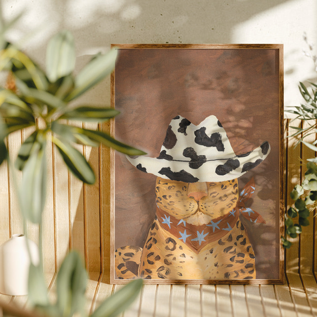 Framed artwork of a leopard wearing a cowboy hat with star patterns, placed on a surface with plants in the background.
