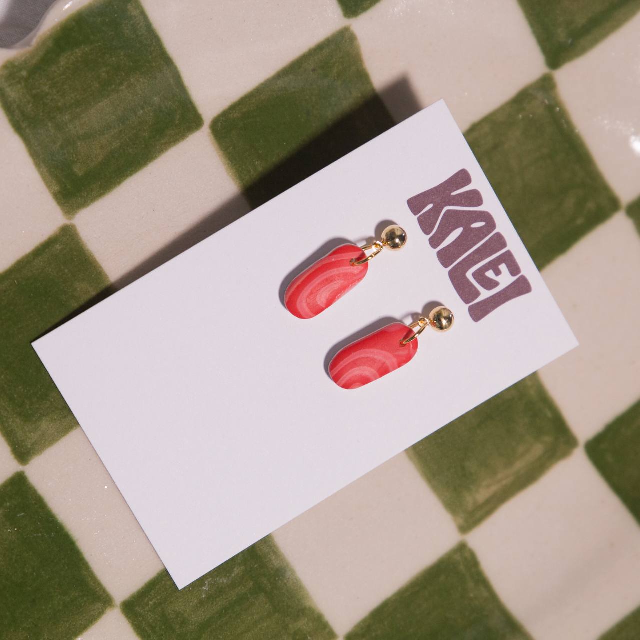 Red earrings on a white card with 'KAEI' branding on a green and white checkered background