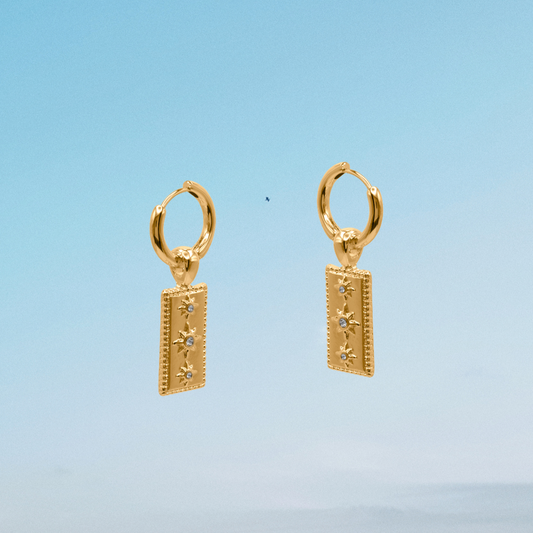 Gold hoop earrings with rectangular pendants against a blue sky and ocean background